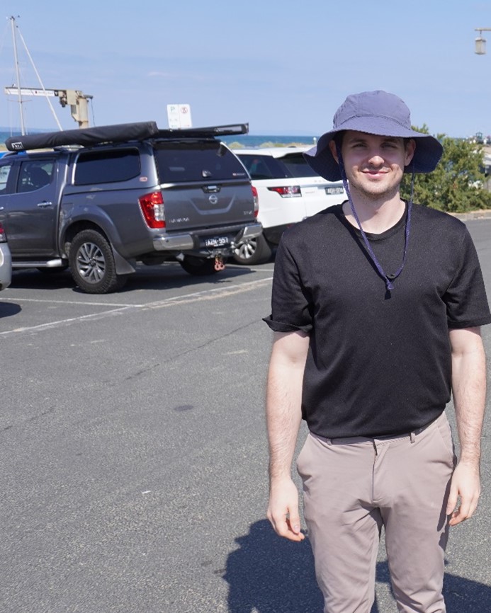 A man is wearing a wide brimmed hat and standing in a car park