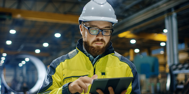 Half upper-body shot of male worker with hard hat looking intently at tablet in an indoor industrial setting